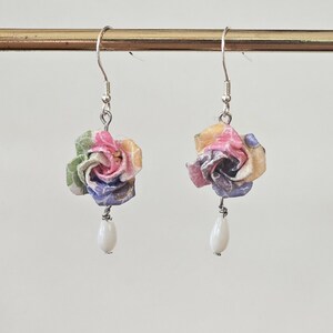 May include: A pair of earrings with paper rose pendants in shades of pink, yellow, green, and purple. Each rose is suspended from a silver hook, with a white, teardrop-shaped bead hanging below. The earrings are handmade and feature a delicate design.