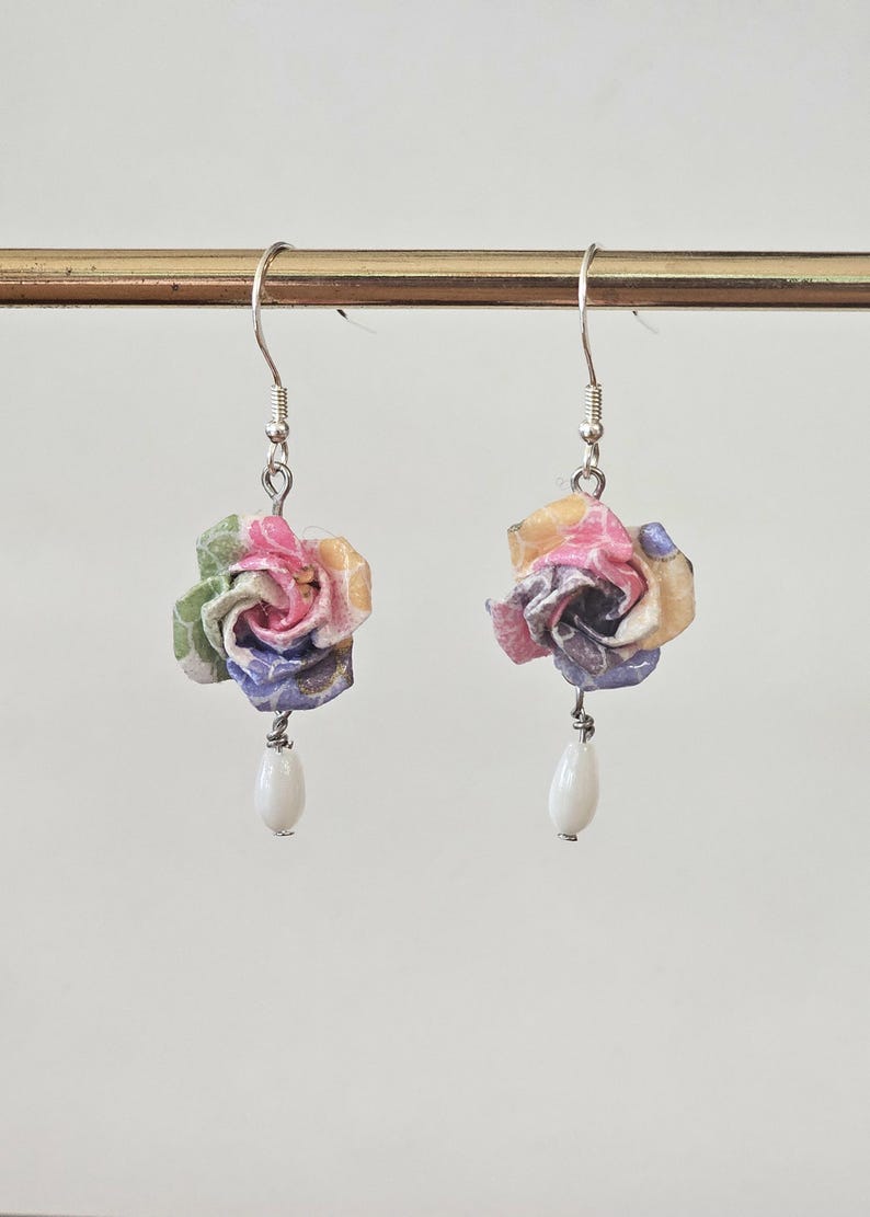 May include: A pair of earrings with paper rose pendants in shades of pink, yellow, green, and purple. Each rose is suspended from a silver hook, with a white, teardrop-shaped bead hanging below. The earrings are handmade and feature a delicate design.