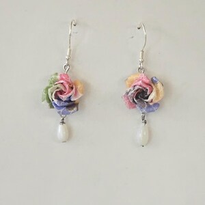 May include: A pair of floral earrings. Each earring has a fabric flower in pink, yellow, blue, and green, with a white teardrop bead hanging below. The earrings have silver hooks. The earrings are handmade.