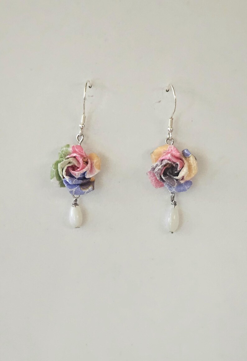 May include: A pair of floral earrings. Each earring has a fabric flower in pink, yellow, blue, and green, with a white teardrop bead hanging below. The earrings have silver hooks. The earrings are handmade.