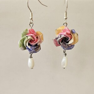 May include: A pair of earrings with paper flower buds in shades of pink, yellow, green, and blue. Each earring has a silver hook and a white, teardrop-shaped bead. The flowers are made from layered paper.