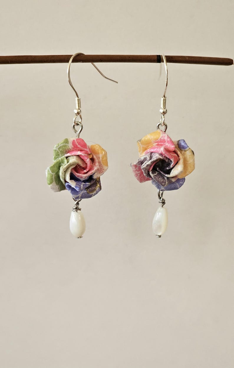May include: A pair of earrings with paper flower buds in shades of pink, yellow, green, and blue. Each earring has a silver hook and a white, teardrop-shaped bead. The flowers are made from layered paper.
