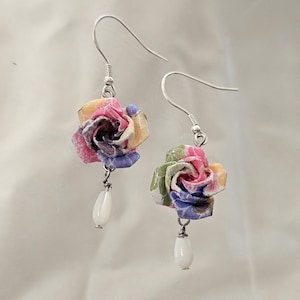 May include: A pair of floral earrings. Each earring features a colourful paper rose in shades of pink, yellow, and blue, with a white teardrop-shaped bead dangling below. The earrings have silver hooks.