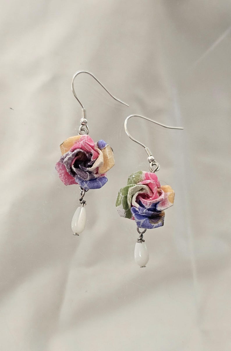 May include: A pair of floral earrings. Each earring features a colourful paper rose in shades of pink, yellow, and blue, with a white teardrop-shaped bead dangling below. The earrings have silver hooks.
