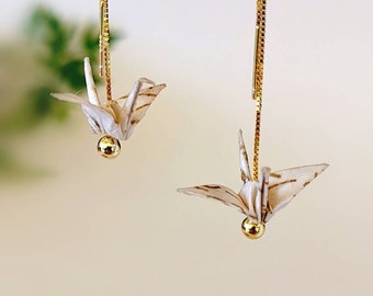 May include: A pair of gold dangle earrings with white origami cranes hanging from them. The cranes have a gold bead at the bottom.