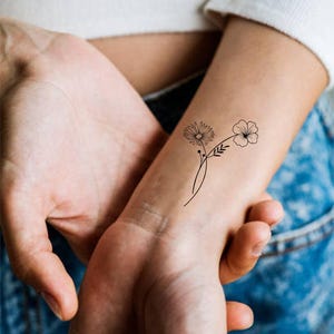 May include: A simple black line drawing of two flowers on a person's wrist. The flowers are stylized and have a delicate, minimalist aesthetic.