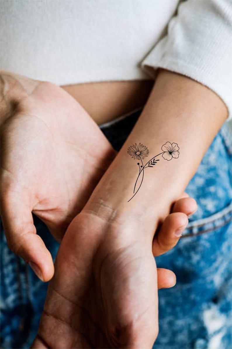 May include: A simple black line drawing of two flowers on a person's wrist. The flowers are stylized and have a delicate, minimalist aesthetic.