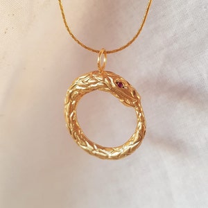 Puede incluir: Un collar de serpiente de oro con un ojo de piedra roja. La serpiente está enroscada en un círculo.