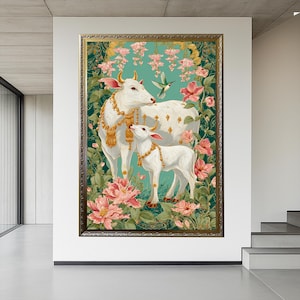 Puede incluir: Una obra de arte enmarcada que presenta dos vacas blancas adornadas con joyas de oro, sobre un fondo de flores rosas y exuberante vegetaci&oacute;n. Un colibr&iacute; revolotea cerca. La obra tiene un fondo turquesa y un marco dorado.