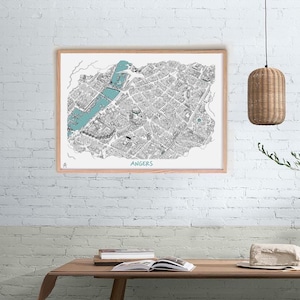 May include: A framed monochrome map of Angers, France, with teal highlighting the river. The map is in a light wood frame and hangs on a white brick wall. The word "ANGERS" is printed in teal at the bottom of the map.
