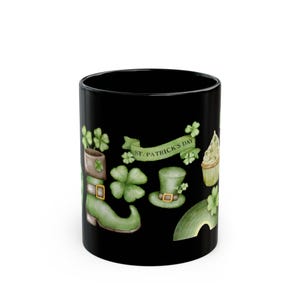 Op de afbeelding: Zwarte keramische mok met illustraties met het thema St. Patrick's Day. Het ontwerp bevat een groene laars, klavers, een kabouterhoed en een banner met de tekst "St. Patrick's Day". Een cupcake is ook afgebeeld.
