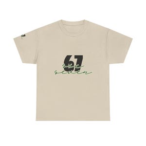 Op de afbeelding: Beige t-shirt met het nummer "67" in zwart en het woord "six seven" in een groen cursief lettertype. Het t-shirt heeft een ronde hals en korte mouwen. De stof lijkt zacht en comfortabel.
