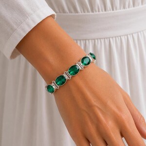 Pode incluir: Uma pulseira de tom prateado com cinco pedras ovais de um verde vibrante e facetadas, dispostas em fila. A pulseira é construída com elos metálicos que conectam cada pedra, criando uma banda contínua que envolve a área do pulso.