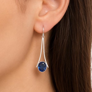 Puede incluir: Pendientes colgantes plateados con una gema ovalada azul oscuro. Los pendientes tienen un diseño geométrico moderno con una forma de triángulo invertido delgada que acuna la gema. Los pendientes se muestran en la oreja de una persona.