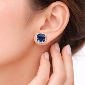 May include: A close-up of a woman's ear with a square, dark blue gemstone stud earring. The earring has a halo of small, clear stones set in silver or white metal. The woman's hand is near her ear, with the earring in focus.