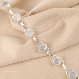 以下が含まれることがあります： 楕円形のオパール宝石をあしらったシルバーブレスレット。宝石はシルバーのリンクと小さな円形コネクタで接続されています。ブレスレットはベージュ色の布の背景に表示されています。