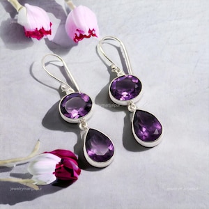 May include: A pair of silver drop earrings featuring oval and teardrop-shaped amethyst gemstones. The earrings have a polished finish and are displayed on a light gray surface with floral accents.