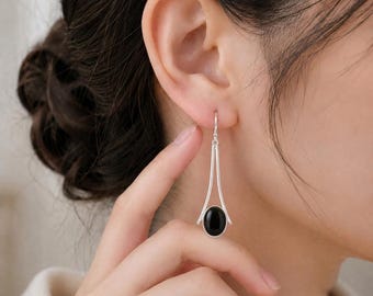 Könnte beinhalten: Ein silberner Ohrhänger mit einem glatten, ovalen schwarzen Stein, der an der Basis eines schlanken, V-förmigen Metallrahmens befestigt ist. Der Ohrring hängt an einem einfachen Haken und besticht durch ein poliertes Metallfinish.