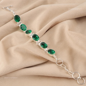 May include: A silver bracelet with oval emerald-green gemstones. The gemstones are set in individual silver bezels and connected by silver links. The bracelet has a toggle clasp and is displayed on a beige fabric.
