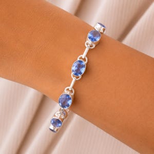 Può includere: Un bracciale a catena color argento caratterizzato da una serie di pietre preziose blu di forma ovale e sfaccettate, incastonate in castoni di metallo lucido. Il bracciale è composto da segmenti collegati, ognuno con una pietra blu traslucida.