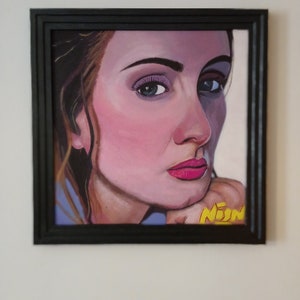 May include: A framed portrait painting of a woman with pink skin tones, blue eyes, and pink lipstick. The painting is in a black frame and features the artist's signature in yellow. The woman is resting her chin on her hand.