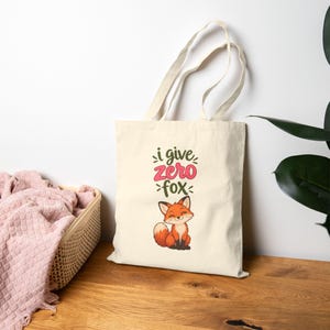 May include: A cream-colored canvas tote bag featuring a cartoon fox illustration and the text "i give zero fox" printed in dark green and pink. The bag has two long shoulder straps and is made of a light-colored fabric with a simple design.