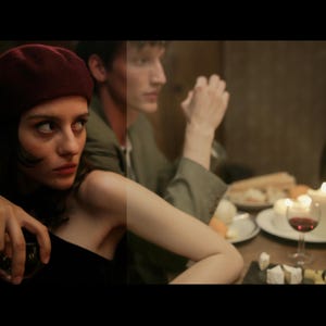 May include: A woman wearing a red beret and a black dress sits at a table with a man. The table is set with wine glasses, bottles of wine, and a plate of food. The woman is looking at the camera.
