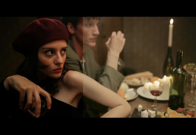 May include: A woman wearing a red beret and a black dress sits at a table with a man. The table is set with wine glasses, bottles of wine, and a plate of food. The woman is looking at the camera.