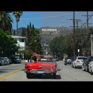 May include: A red vintage convertible car drives down a street lined with parked cars and palm trees. The Hollywood sign is visible on a distant hillside under a blue sky.