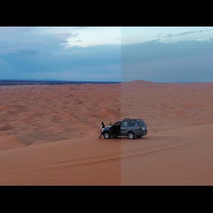 以下が含まれることがあります： 砂丘が広がる砂漠の風景を空撮。曇り空の下、濃いグレーのSUVが砂丘に駐車し、近くに人が立っています。砂は暖かく赤茶色です。