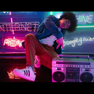 May include: A person in a blue jacket and tan pants poses next to a silver boombox. The background features neon signs that read "INTERNET" and "RENT". The person is wearing white sneakers with black stripes.