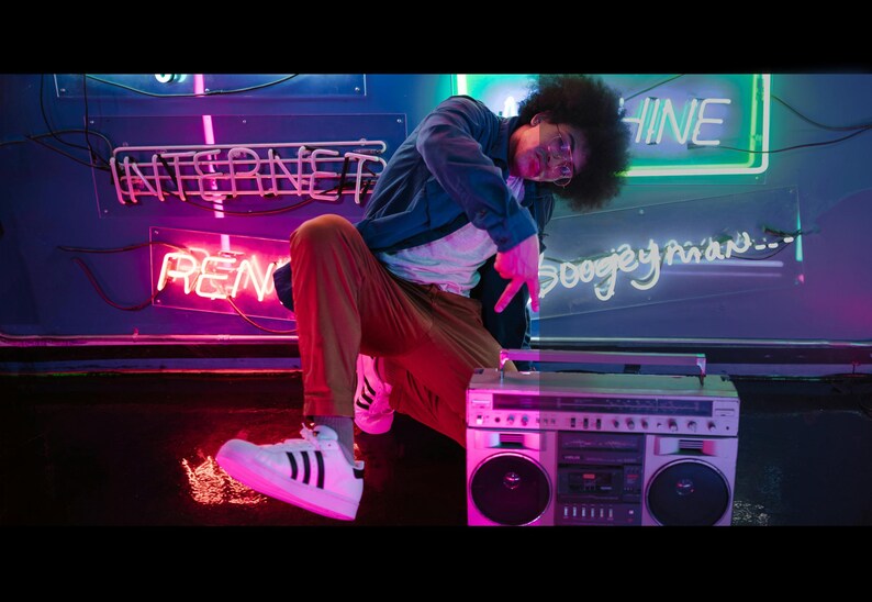 May include: A person in a blue jacket and tan pants poses next to a silver boombox. The background features neon signs that read "INTERNET" and "RENT". The person is wearing white sneakers with black stripes.