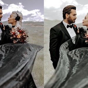 May include: A couple in black wedding attire stand in a field, the bride holding a bouquet of red and orange flowers. The bride's veil is flowing in the wind.