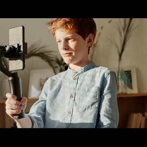 May include: A young person with red hair is holding a smartphone attached to a black stabilizer. The person is wearing a light blue button-down shirt.