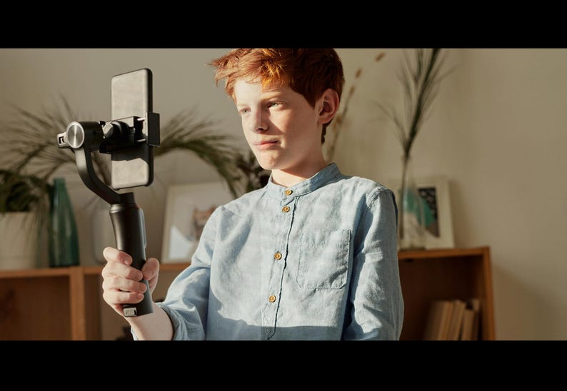 May include: A young person with red hair is holding a smartphone attached to a black stabilizer. The person is wearing a light blue button-down shirt.