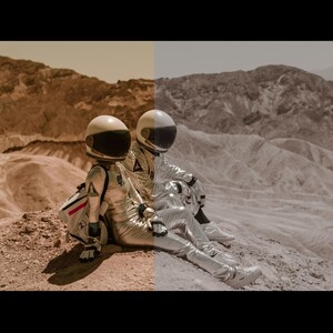 May include: Two astronauts in silver spacesuits sit on a rocky, red-brown landscape. The astronauts are wearing helmets and are looking out at the view.
