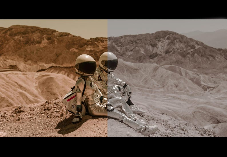 May include: Two astronauts in silver spacesuits sit on a rocky, red-brown landscape. The astronauts are wearing helmets and are looking out at the view.