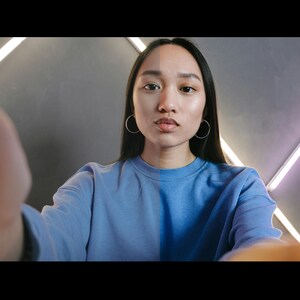 May include: A woman with long black hair wearing a light blue sweatshirt looks directly at the camera. The background is a gray wall with white diagonal lines.
