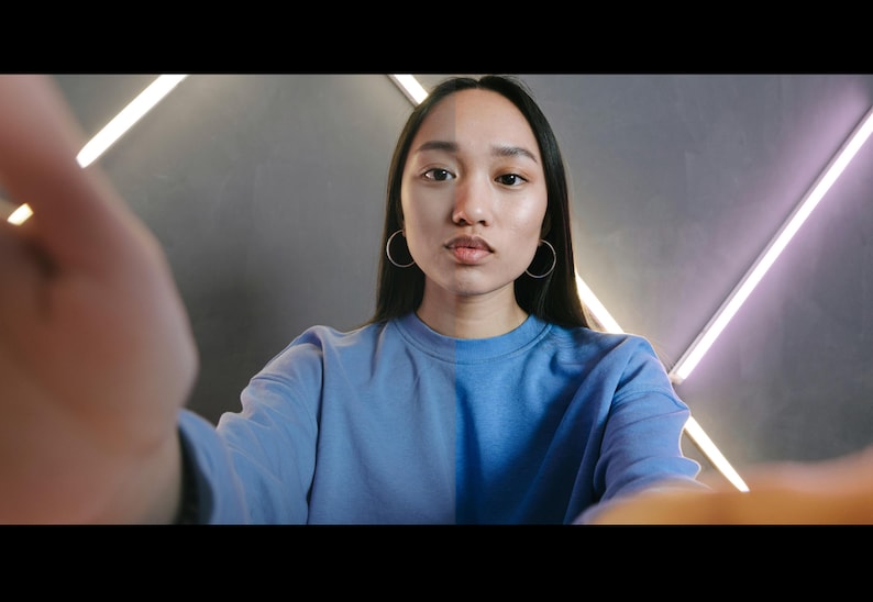 May include: A woman with long black hair wearing a light blue sweatshirt looks directly at the camera. The background is a gray wall with white diagonal lines.