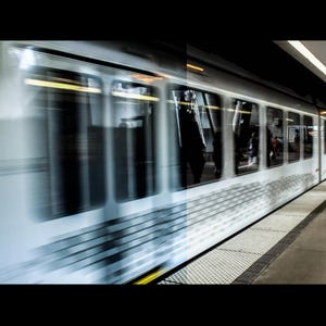 Puede incluir: Un tren en movimiento r&aacute;pido en una estaci&oacute;n de metro. El tren es blanco y plateado con ventanas, y est&aacute; borroso debido a su movimiento. El and&eacute;n es gris con una superficie texturizada, y una persona est&aacute; de pie en el and&eacute;n.