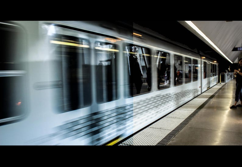 Puede incluir: Un tren en movimiento r&aacute;pido en una estaci&oacute;n de metro. El tren es blanco y plateado con ventanas, y est&aacute; borroso debido a su movimiento. El and&eacute;n es gris con una superficie texturizada, y una persona est&aacute; de pie en el and&eacute;n.