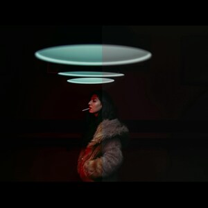 May include: A woman in a brown and black fur coat stands in a dark room with a series of glowing, circular lights above her head.