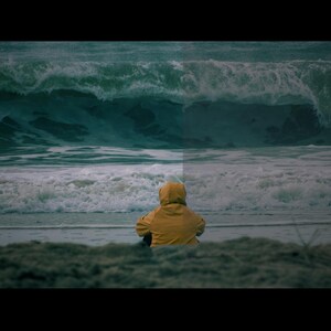 以下が含まれることがあります： 黄色いフード付きジャケットを着た人が、海を眺めながら砂浜に座っています。背景には大きな波が打ち寄せ、ドラマチックなシーンを作り出しています。全体的な色調は落ち着いており、緑、青、黄色の色合いが使われています。