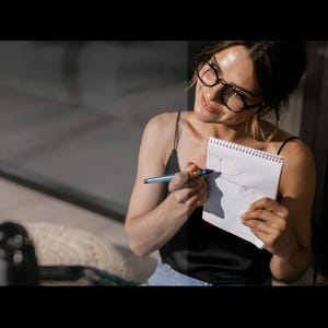 May include: A woman wearing glasses and a black tank top is drawing on a white notebook with a blue pen. She is smiling and looking at the notebook.