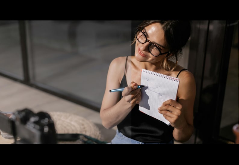 May include: A woman wearing glasses and a black tank top is drawing on a white notebook with a blue pen. She is smiling and looking at the notebook.