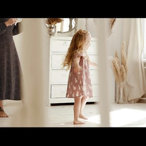 May include: A young girl in a pink dress with white animal print is standing in a room with white walls and a white dresser. She is smiling and looking to the right.