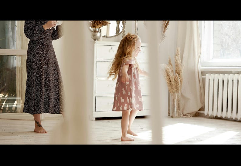 May include: A young girl in a pink dress with white animal print is standing in a room with white walls and a white dresser. She is smiling and looking to the right.