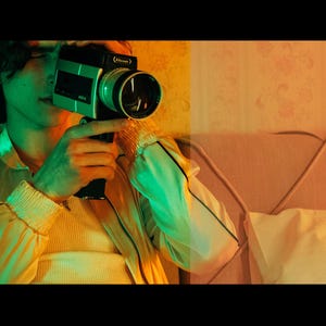 以下が含まれることがあります： 大きなレンズが付いた、緑と黒のヴィンテージSuper 8フィルムカメラを持っている人。その人は黒いトリムが付いた黄色のジャケットを着ています。シーンは緑とオレンジ色の光で照らされています。