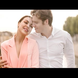 May include: A couple standing close together, looking at each other with smiles on their faces. The woman is wearing a pink blazer and the man is wearing a white button-down shirt. They are standing in front of a city background.