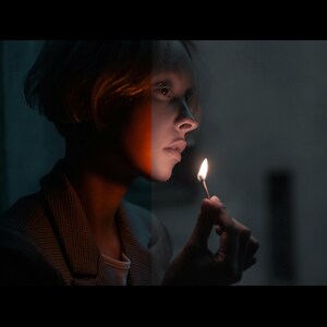 May include: A person with short, light brown hair holds a lit match in their right hand. The person is looking to the right of the frame. The person's face is partially obscured by a shadow, but the left side of their face is painted with a white and orange stripe.